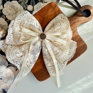 Op de afbeelding: Een crèmekleurige kanten strik met een decoratief midden, geplaatst op een houten snijplank. De strik heeft delicate kanten details en lange, crèmekleurige linten. De achtergrond bevat een bloemenpatroon.