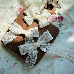 May include: Close-up of several cream lace bows and ribbon on a wooden chopping board. The bows have a delicate floral pattern and are arranged on floral fabric. A dark brown leather strap is attached to the chopping board.