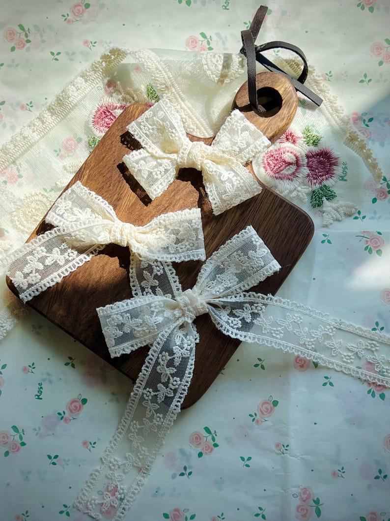 Puede incluir: Primer plano de varios lazos y cintas de encaje color marfil sobre una tabla de cortar de madera. Los lazos tienen un delicado estampado floral y est&aacute;n dispuestos sobre una tela estampada con flores. Una correa de cuero marr&oacute;n oscuro est&aacute; sujeta a la tabla.