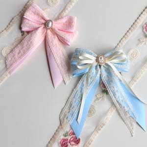 May include: Two decorative hair bows. One is light pink with a pearl-like center and lace trim. The other is light blue with a cream-colored center and lace trim. Both bows have long, flowing ribbons.