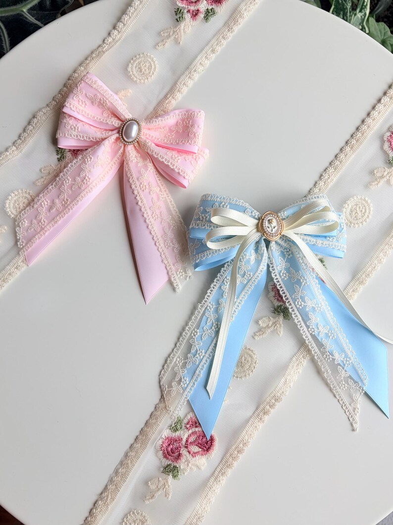 May include: Two decorative hair bows. One is light pink with a pearl-like center and lace trim. The other is light blue with a cream-colored center and lace trim. Both bows have long, flowing ribbons.