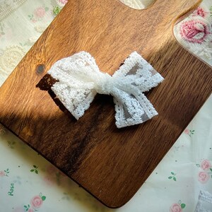 Puede incluir: Un lazo blanco de encaje, probablemente para el cabello o decoraci&oacute;n, descansa sobre una tabla de cortar de madera. El lazo est&aacute; hecho de encaje delicado de tejido abierto, y la madera tiene un tono c&aacute;lido. El fondo incluye tela floral.