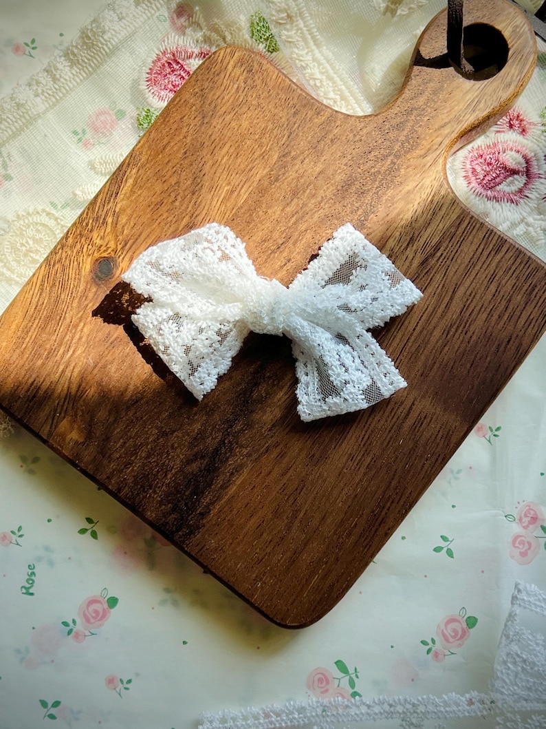 Puede incluir: Un lazo blanco de encaje, probablemente para el cabello o decoraci&oacute;n, descansa sobre una tabla de cortar de madera. El lazo est&aacute; hecho de encaje delicado de tejido abierto, y la madera tiene un tono c&aacute;lido. El fondo incluye tela floral.