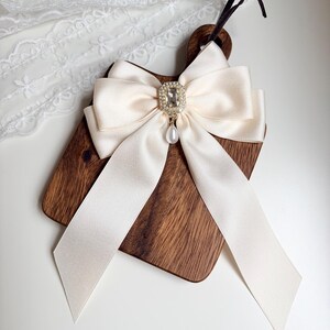 May include: A decorative wooden cutting board with a cream-colored satin bow. The bow features a square rhinestone embellishment with a dangling pearl. The board has a dark brown finish and a handle.