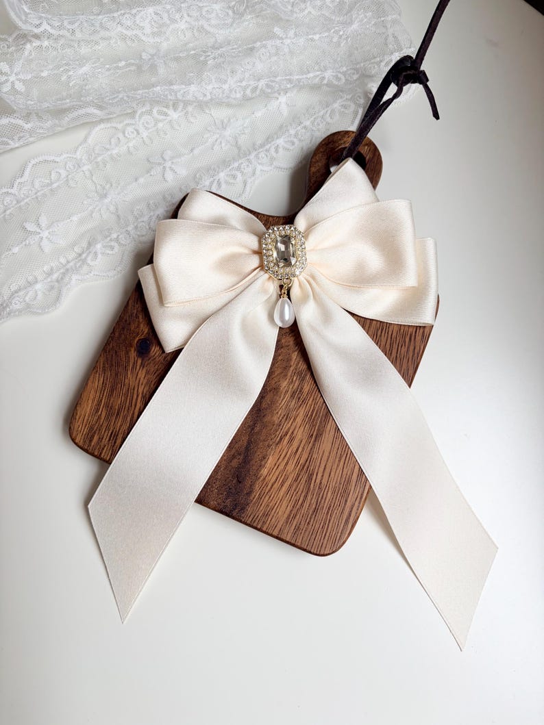 May include: A decorative wooden cutting board with a cream-colored satin bow. The bow features a square rhinestone embellishment with a dangling pearl. The board has a dark brown finish and a handle.