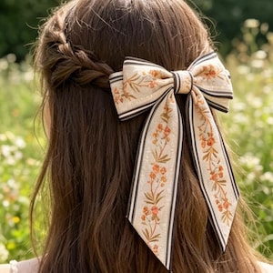 以下が含まれることがあります： 茶色の髪に、花柄と黒いストライプが入ったクリーム色のヘアリボンが付いています。リボンは、長く流れるリボンで大きなリボンの形に結ばれています。花柄にはオレンジとグリーンのアクセントが含まれています。