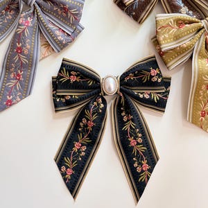 May include: Several decorative hair bows in various colors, including black, gray, brown, and gold. The bows feature floral embroidery and are accented with gold trim. The central bow has a pearl-like embellishment.