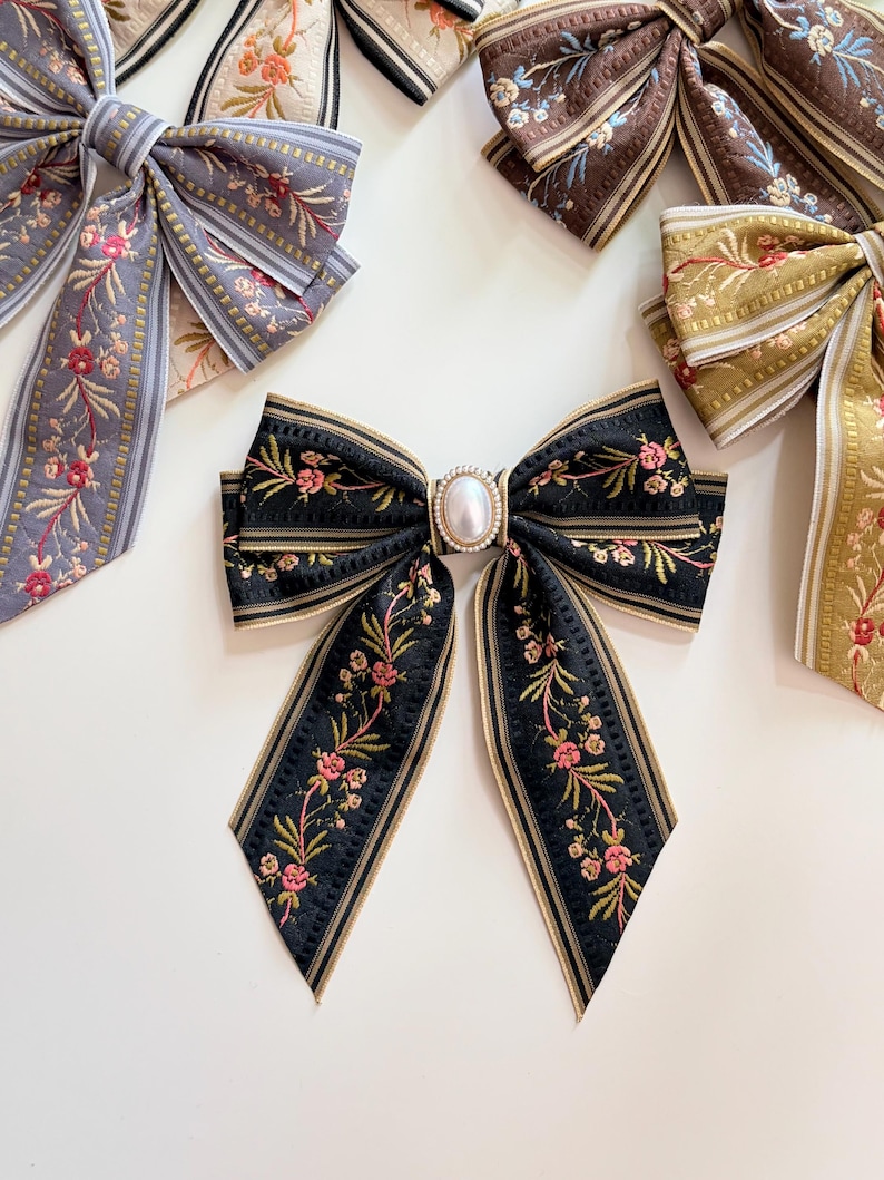 May include: Several decorative hair bows in various colors, including black, gray, brown, and gold. The bows feature floral embroidery and are accented with gold trim. The central bow has a pearl-like embellishment.