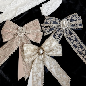 Peut inclure: Plusieurs nœuds décoratifs avec des détails de dentelle et des embellissements. Les nœuds sont de différentes couleurs, notamment rose blush, bleu marine et ivoire, avec des accents de strass et de perles scintillantes. Présentés sur un fond noir.