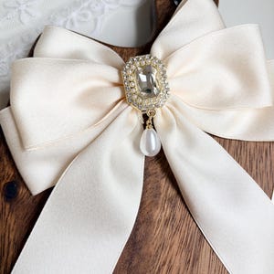 May include: Cream-colored satin bow with a gold and rhinestone centerpiece and a dangling pearl. The bow is set against a wooden background, creating a contrast of textures and colors. The bow's elegant design suggests it could be used as a hair accessory or decorative element.
