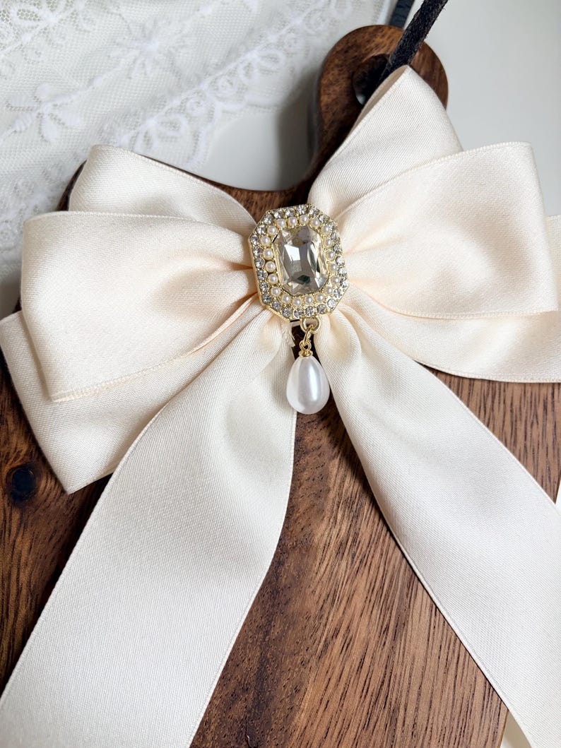 May include: Cream-colored satin bow with a gold and rhinestone centerpiece and a dangling pearl. The bow is set against a wooden background, creating a contrast of textures and colors. The bow's elegant design suggests it could be used as a hair accessory or decorative element.