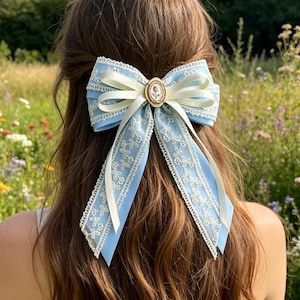 May include: A light blue hair bow with white lace trim and a cream-colored ribbon. The bow features a gold-colored circular centerpiece with a cameo design. The bow is worn in the hair.