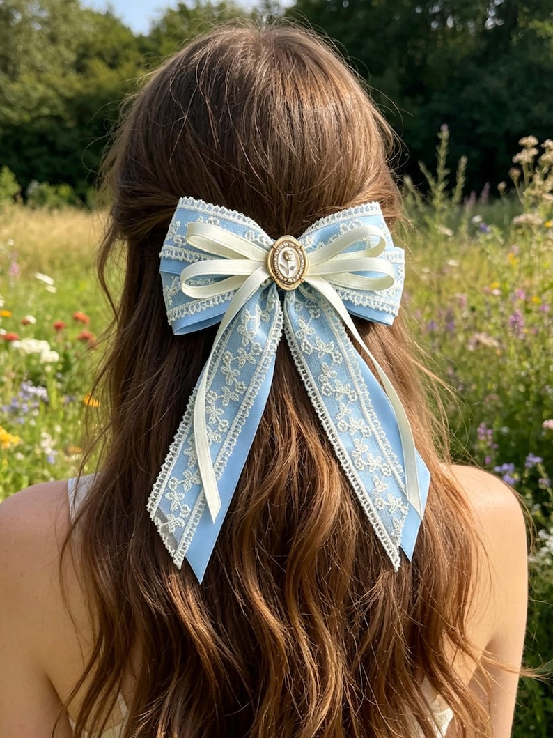May include: A light blue hair bow with white lace trim and a cream-colored ribbon. The bow features a gold-colored circular centerpiece with a cameo design. The bow is worn in the hair.