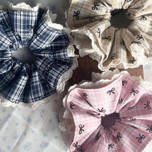 May include: A close-up of three fabric scrunchies. One is navy and white plaid, another is tan plaid with black bow details, and the third is pink plaid with black bow details. Each has a white lace trim.