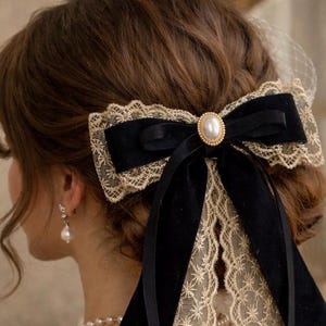 Könnte beinhalten: Ein schwarzes Haaraccessoire mit Samtschleife, goldfarbener Spitzentrimmung und einer Perle in der Mitte. Die Schleife ist an einer Haarspange befestigt und wird im Haar getragen. Das Accessoire ist im Vintage-Stil gehalten und eignet sich für besondere Anlässe.
