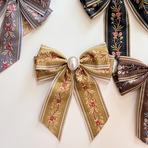 May include: An assortment of decorative fabric bows in various colors. The bows feature floral embroidery and striped edges. One bow has a pearl-like embellishment in the center. Colors include gold, black, brown, and gray.