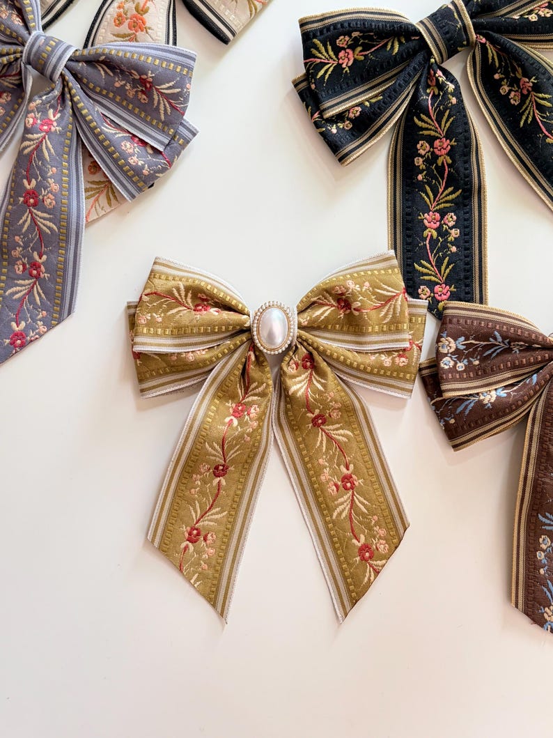 May include: An assortment of decorative fabric bows in various colors. The bows feature floral embroidery and striped edges. One bow has a pearl-like embellishment in the center. Colors include gold, black, brown, and gray.