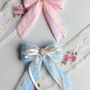 May include: Two decorative hair bows. One is light pink with a pearl-centered embellishment, and the other is light blue with a cameo-style center. Both bows feature lace trim and long, flowing ribbons.