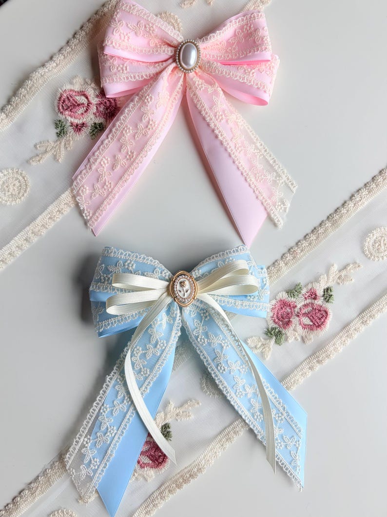 May include: Two decorative hair bows. One is light pink with a pearl-centered embellishment, and the other is light blue with a cameo-style center. Both bows feature lace trim and long, flowing ribbons.