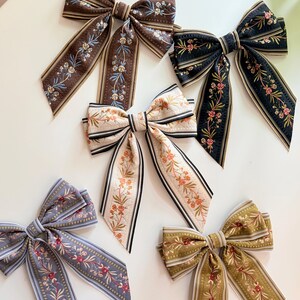 May include: An assortment of decorative hair bows in various colors, including brown, black, cream, gray, and olive green. Each bow features a floral pattern with coordinating striped edges. The bows are arranged on a white surface.
