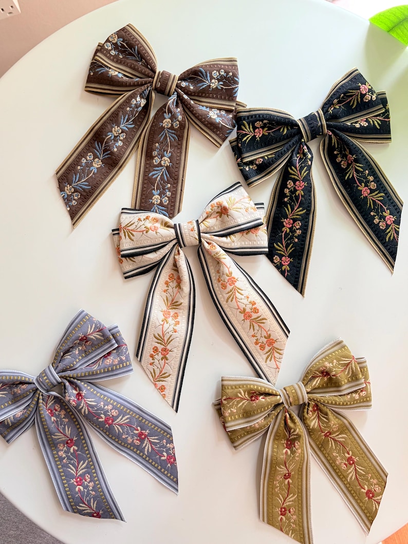 May include: An assortment of decorative hair bows in various colors, including brown, black, cream, gray, and olive green. Each bow features a floral pattern with coordinating striped edges. The bows are arranged on a white surface.