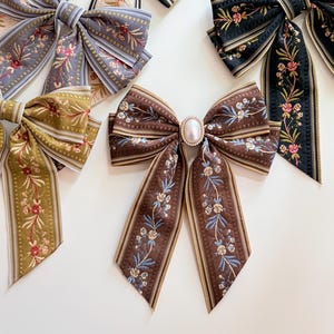 May include: Several decorative hair bows in various colors and patterns. The bows feature floral designs and striped ribbons. One bow has a pearl-like embellishment in the center. Colors include brown, black, gray, and olive green.