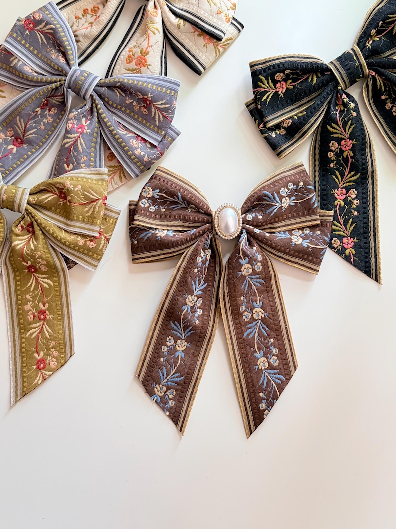 May include: Several decorative hair bows in various colors and patterns. The bows feature floral designs and striped ribbons. One bow has a pearl-like embellishment in the center. Colors include brown, black, gray, and olive green.
