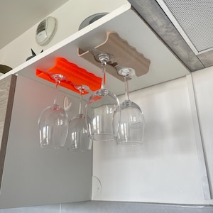 Peut inclure: Un meuble de cuisine avec deux supports à verres à vin. Un support est orange, l'autre est de couleur brun clair. Quatre verres à vin transparents sont suspendus aux supports.