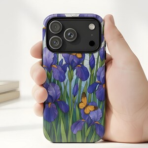 May include: A smartphone case featuring a watercolor design of purple irises and a small orange butterfly. The case has a black camera module and is held in a hand. The background includes a vase of irises and a stack of books.