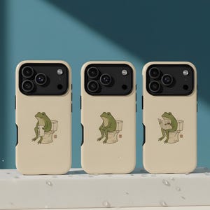 May include: Three phone cases with a cream-colored background and a humorous illustration of a green frog on a toilet. The frog is depicted in different poses, including reading a newspaper. The cases have a black camera module.