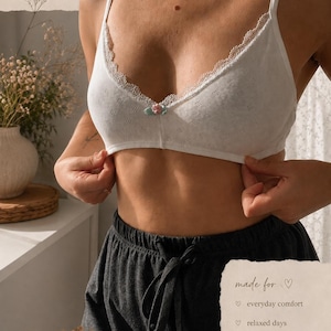 Könnte beinhalten: Ein weißes Bralette aus strukturiertem Stoff mit dünnen Trägern und zarter Spitzenborte am Ausschnitt. Ein kleiner rosa-grüner Blumenakzent befindet sich mittig an der Brust. Der Text 100% cotton soft & breathable, made for everyday comfort, relaxed days, natural feeling, light support, and natural fit ist sichtbar.
