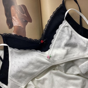 以下が含まれることがあります： 黒と白の2つのブラジャーが表示されています。黒いブラジャーはレースのトリムと小さな赤いバラのアクセントが付いています。白いブラジャーはドット柄とピンクのバラのアクセントが特徴です。どちらのブラジャーも細いストラップが付いています。