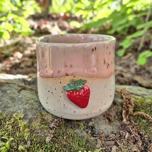 May include: A handmade ceramic cup with a strawberry design. The cup has a light pink top and a speckled beige bottom. A red strawberry with green leaves is attached to the front of the cup.