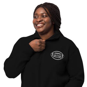 Peut inclure: Un sweat-shirt noir à fermeture éclair quart de col avec un motif brodé blanc sur la poitrine gauche. Le motif représente un ballon de football américain avec le mot MOM au centre, entouré de coutures blanches imitant les lacets.