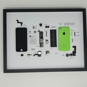 Op de afbeelding: Een ingelijste afbeelding met de gedemonteerde onderdelen van een iPhone 5C. De afbeelding toont een zwarte telefoon, een groene telefoon en diverse interne onderdelen, allemaal gelabeld. De lijst is zwart en de achtergrond is wit.
