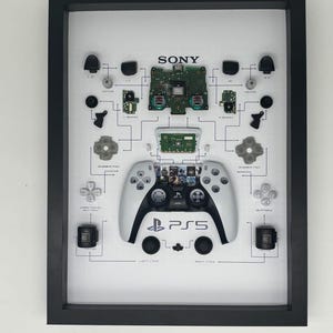 Könnte beinhalten: Gerahmtes Kunstwerk mit einem zerlegten PlayStation 5-Controller. Der schwarz-weiße Controller ist von seinen Komponenten umgeben, die mit Text beschriftet sind. Das Sony-Logo und das PS5-Logo sind sichtbar.