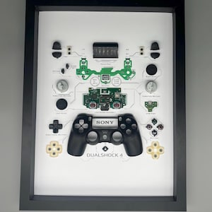 Op de afbeelding: Een ingelijste, gedemonteerde PlayStation DualShock 4-controller. Het zwarte frame omsluit een witte achtergrond met de componenten van de controller, waaronder de printplaat, knoppen en joysticks. De behuizing van de controller staat in het midden, met het "SONY"-logo.