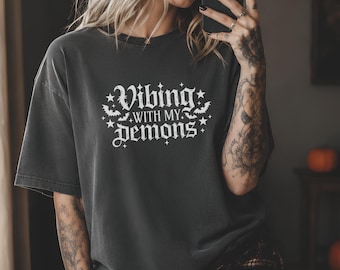 Op de afbeelding: Een antracietgrijs T-shirt met korte mouwen en witte tekst "Vibing with my demons", omringd door kleine ster- en vleermuisafbeeldingen. Het T-shirt heeft een ronde hals en een relaxte pasvorm, met de afbeelding gecentreerd op de borst.
