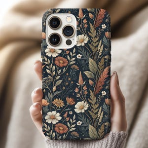 K&ouml;nnte beinhalten: Eine dunkelmarineblaue Smartphone-H&uuml;lle mit einem wiederkehrenden Muster aus Wildblumen, darunter wei&szlig;e G&auml;nsebl&uuml;mchen, rostfarbene Bl&uuml;ten sowie verschiedene gr&uuml;ne und braune Bl&auml;tter. Die H&uuml;lle hat eine Aussparung f&uuml;r ein Dreifach-Kamerasystem.