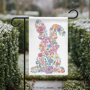 May include: A garden flag with a floral bunny design. The bunny is made of colorful flowers, including roses and tulips, with a pink bow. The flag is displayed on a black metal stand, ideal for spring or Easter.