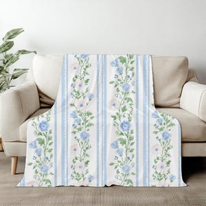 May include: A white throw blanket with a floral pattern of blue and pink flowers and green leaves. The design is arranged in vertical stripes with light blue stripes. The blanket is draped over a beige sofa.