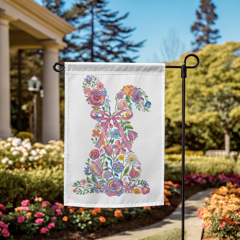 May include: A white garden flag with a floral bunny design. The bunny is made of colourful flowers and a pink ribbon. The flag is displayed outdoors on a black metal stand, surrounded by greenery and flowers.