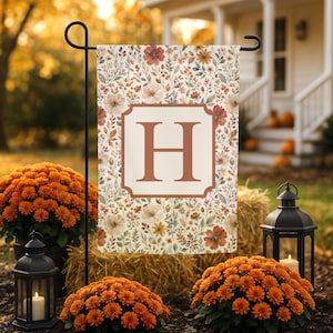 Puede incluir: Una bandera de jardín rectangular con un patrón floral en tonos naranja, marrón y crema. El centro muestra una letra H grande y en negrita con una fuente de color óxido, encerrada dentro de un borde decorativo sobre un fondo crema claro.
