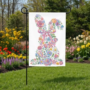 May include: A white garden flag with a floral bunny design. The bunny is made of colourful flowers and a pink ribbon bow. The flag is displayed on a black metal stand in a garden setting.