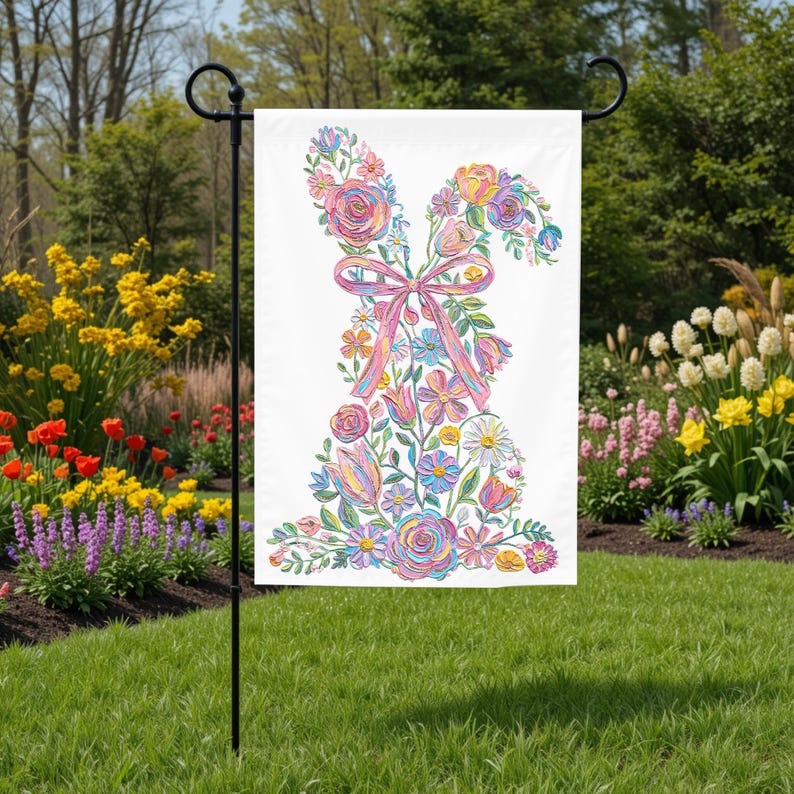 May include: A white garden flag with a floral bunny design. The bunny is made of colourful flowers and a pink ribbon bow. The flag is displayed on a black metal stand in a garden setting.