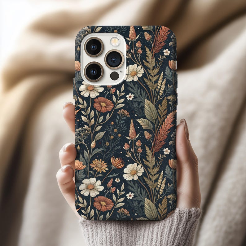 K&ouml;nnte beinhalten: Eine dunkelmarineblaue Smartphone-H&uuml;lle mit einem wiederkehrenden Muster aus Wildblumen, darunter wei&szlig;e G&auml;nsebl&uuml;mchen, rostfarbene Bl&uuml;ten sowie verschiedene gr&uuml;ne und braune Bl&auml;tter. Die H&uuml;lle hat eine Aussparung f&uuml;r ein Dreifach-Kamerasystem.