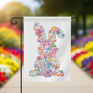May include: A white garden flag with a floral bunny design. The bunny is made up of colourful flowers, including roses, daisies, and tulips, with a pink ribbon bow. The flag is displayed on a black metal stand in a garden setting.