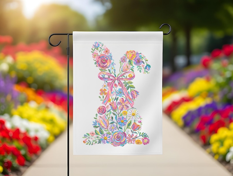 May include: A white garden flag with a floral bunny design. The bunny is made up of colourful flowers, including roses, daisies, and tulips, with a pink ribbon bow. The flag is displayed on a black metal stand in a garden setting.