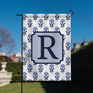 May include: A garden flag featuring a floral pattern with blue flowers on a white background. The flag has a light blue square with the letter "R" in a darker blue. The flag is hanging on a black metal pole.