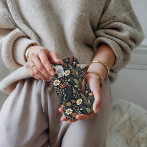 K&ouml;nnte beinhalten: Eine rechteckige Smartphone-H&uuml;lle mit dunklem Hintergrund und einem dichten, komplizierten Muster aus verschiedenen Wildblumen, Gr&auml;sern und Bl&auml;ttern in Creme-, gedeckten Orange-, Braun- und Salbeigr&uuml;nt&ouml;nen, mit Aussparung f&uuml;r die Kamera.